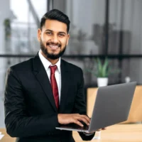 portrait-successful-confident-elegant-indian-arabian-young-businessman-suit-holding-open-laptop-his-hand-stand-near-desktop-his-modern-office-looking-camera-smiling_754108-631